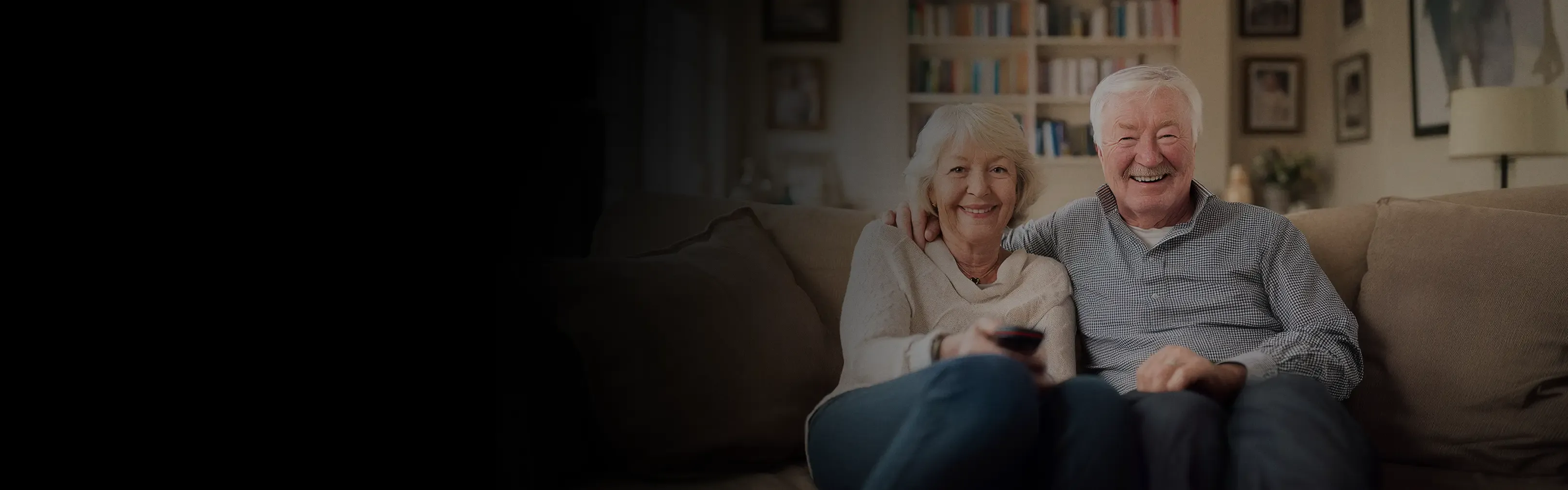 An older couple sits smiling on a couch, the woman holding a remote control.